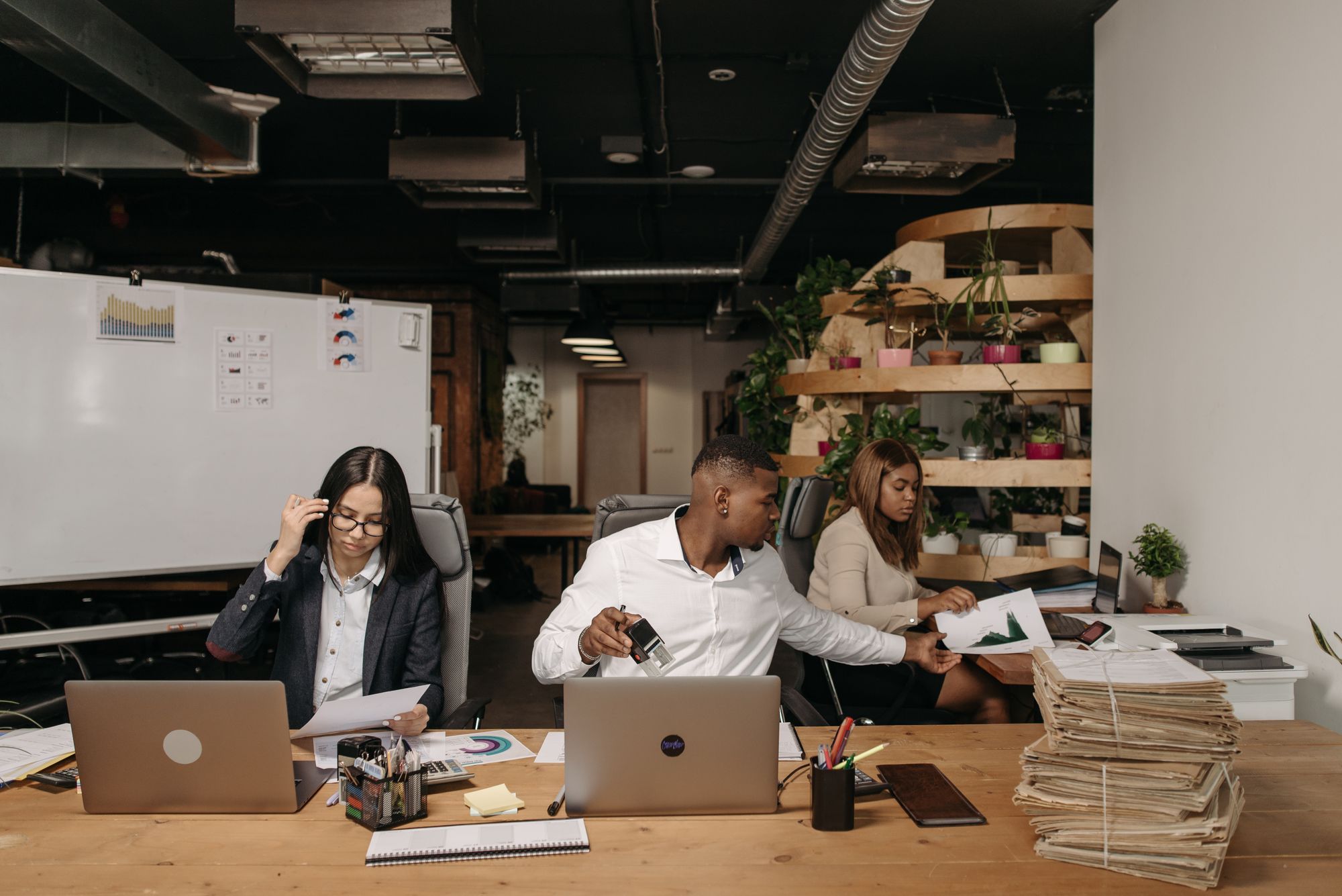 Três pessoas revisam papéis e anotações em uma mesa de escritório com notebooks.